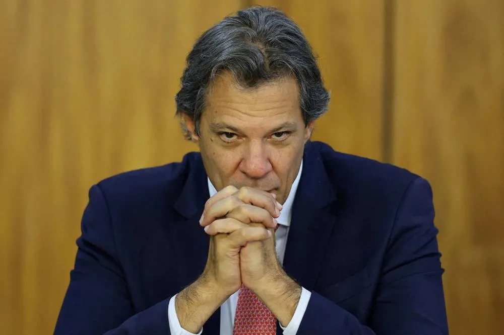 Um homem com cabelo grisalho e liso, vestindo um terno escuro e uma gravata vermelha, está sentado com as mãos unidas em frente ao rosto, em uma expressão pensativa. Ao fundo, há uma parede de madeira clara. O ministro da Fazenda, Fernando Haddad, em Bras