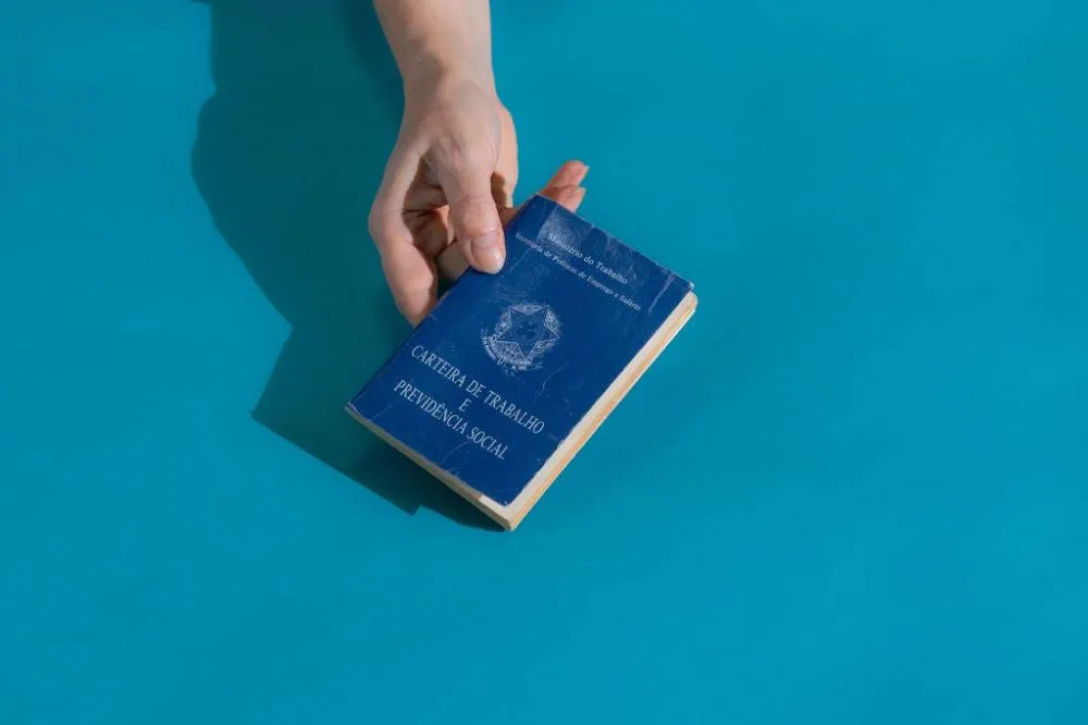 Imagem mostra mão segurando uma carteira de trabalho sobre uma superfície azul Imagem da CLT; IBGE investiga mercado de trabalho formal e informal no Brasil - Gabriel Cabral - 4.out.23/Folhapress